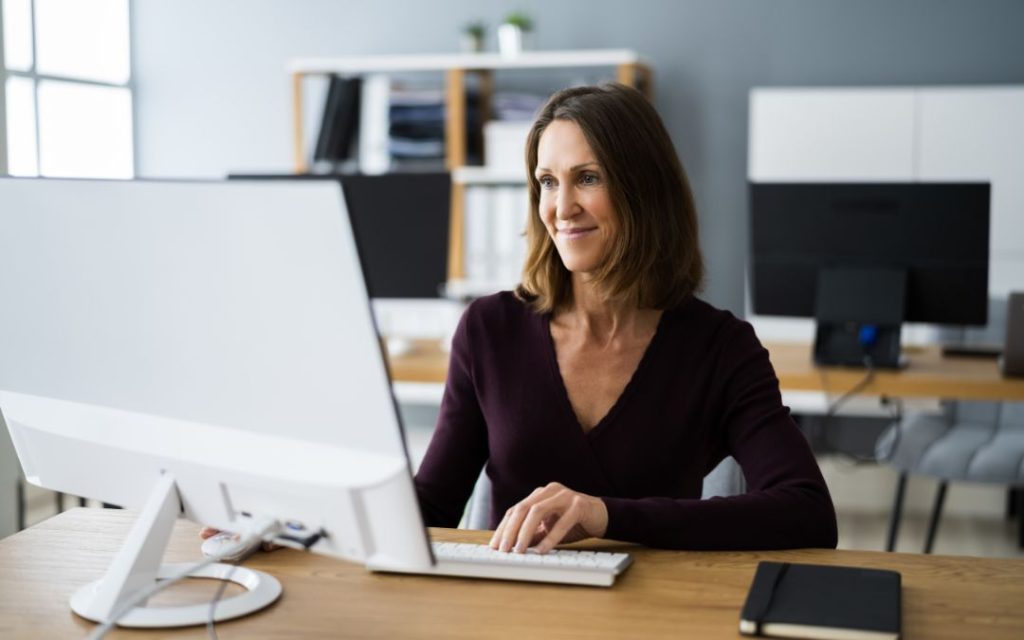 Eine Frau sitzt an einem hellen Holztisch, auf dem ein großer weißer Monitor steht und ein schwarzes Notebook liegt; im Hintergrund stehen Tische, auf denen große schwarze Monitore stehen