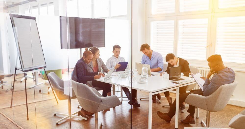 Przeszklona sala, w której odbywa się spotkanie – flipchart, ekran, stół i kilka osób siedzących z laptopami 
